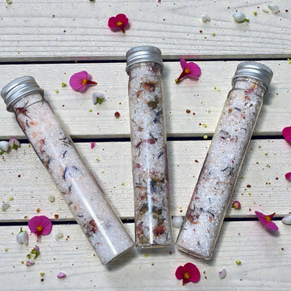 Three glass tubes filled with decorative salt mixtures on a wooden surface with scattered flowers.